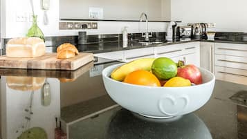 Una cocina con encimera negra, un bol blanco lleno de fruta, una hogaza de pan y una tabla de cortar con una baguette.