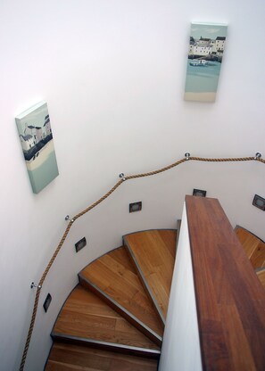 A spiral staircase with wooden steps and a rope handrail, leading to a landing with a framed artwork on the wall. - Riviera Apartments (Teignmouth)