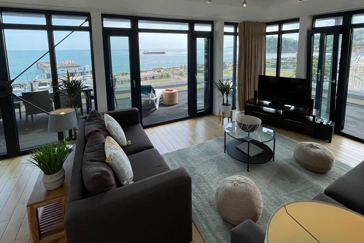 Un salon spacieux avec un grand canapé, une table basse en verre et un téléviseur à écran plat. La pièce est dotée de grandes fenêtres offrant une vue sur la mer et une jetée.