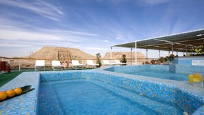 Piscina com azulejos de mosaico azul, cercada por espreguiçadeiras e uma área coberta com vista para uma colina arenosa.