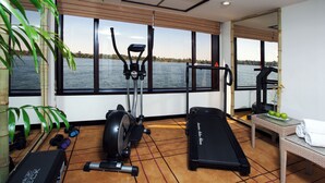Academia com equipamentos de ginástica, esteira e vista para a água através de grandes janelas.