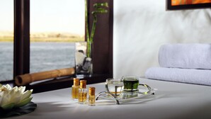 Ambiente de spa com uma flor de lótus, frascos de óleos essenciais e uma bandeja com um copo de água e uma bebida verde, visto por uma janela com vista para um corpo d'água.