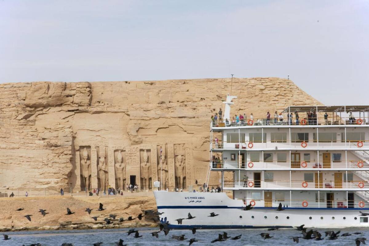 A cruise ship with multiple decks and lifebuoys, docked near a large rock formation with ancient carvings.
