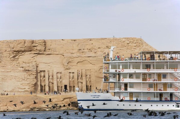 A cruise ship with multiple decks and lifebuoys, docked near a large rock formation with ancient carvings. -  Mövenpick Prince Abbas Lake Nasser Cruise, Every Monday Four Nights From Aswan, Every Friday Three nights From Abu-Simbel - Including Excursions and Sightseeing (Aswan)