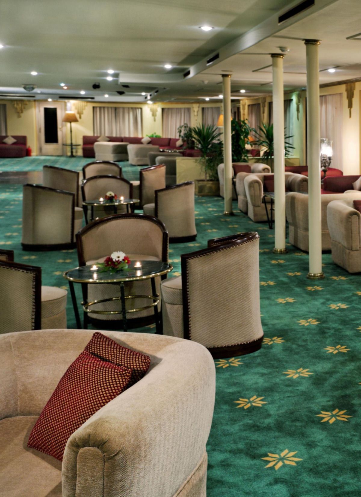 A hotel lobby with a green carpet, multiple seating areas, and a central round table with a flower arrangement.