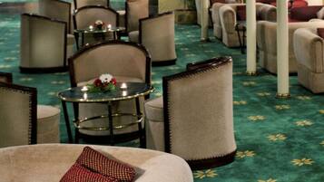 A hotel lobby with a green carpet, multiple seating areas, and a central round table with a flower arrangement.