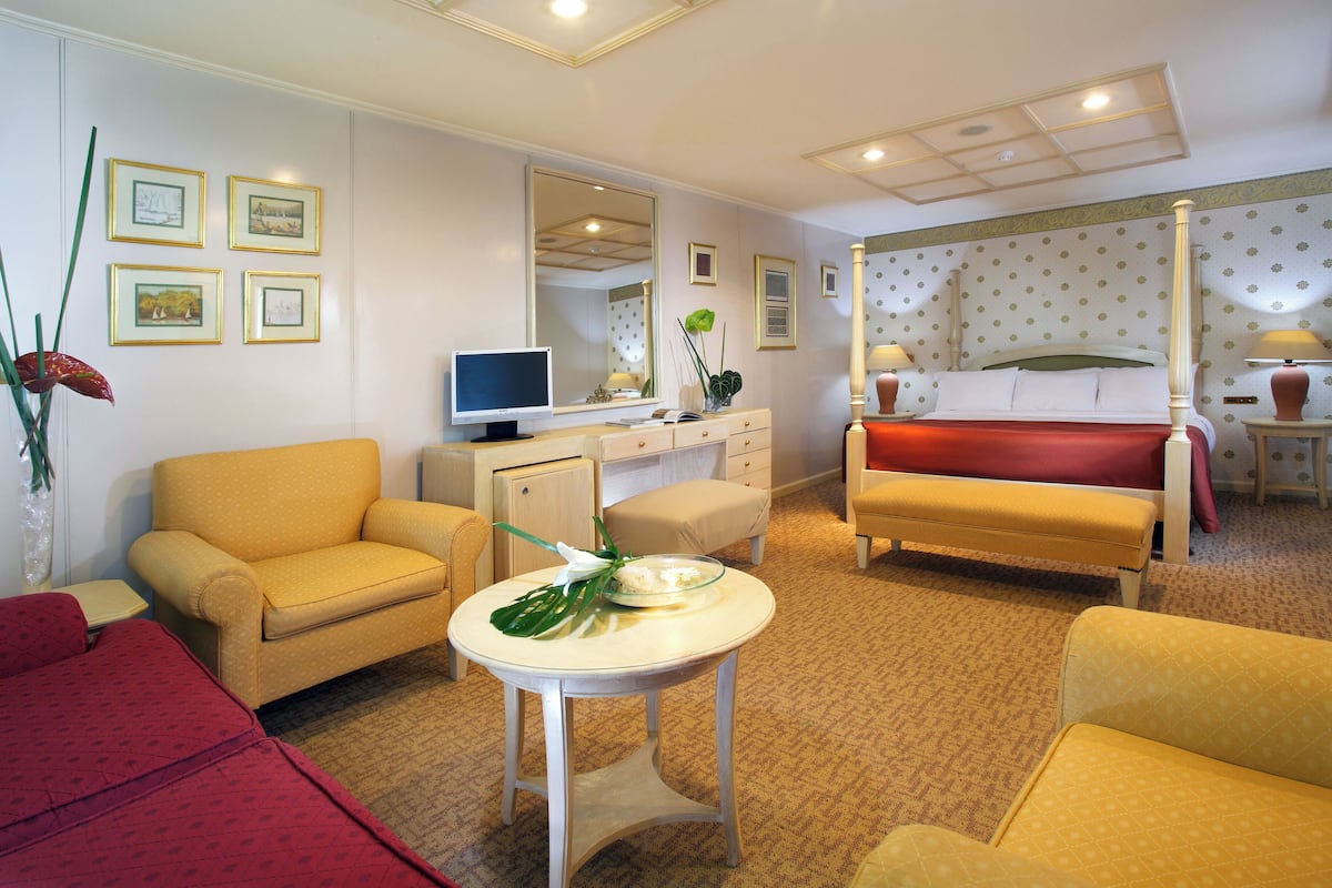 A hotel room with a yellow sofa, a red sofa, a small table with a vase and plant, a bed with a red bedspread, a television, and a mirror.