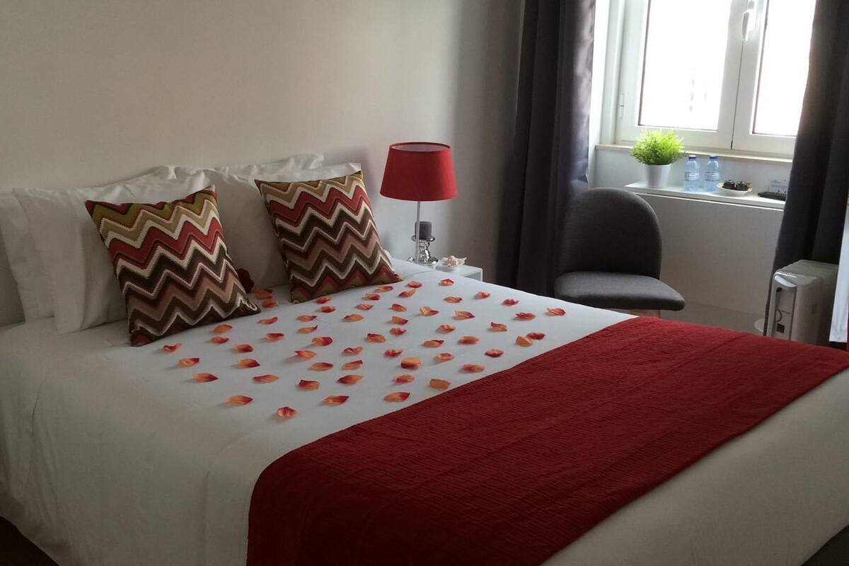 Una cama bien hecha con una colcha roja, dos almohadas estampadas y pétalos de rosa esparcidos sobre la cama.