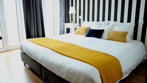 A neatly made bed with a white and yellow bedspread, a gray upholstered headboard, and a bedside table with a lamp. - Aveiro White House (Aveiro)