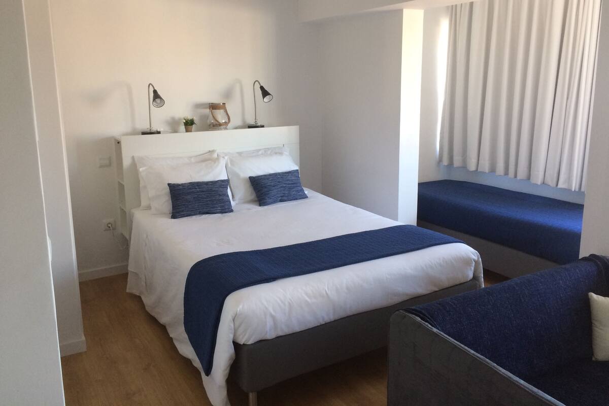 Un dormitorio con cama, mesitas de noche, lámparas de noche, una ventana con cortinas y una silla.