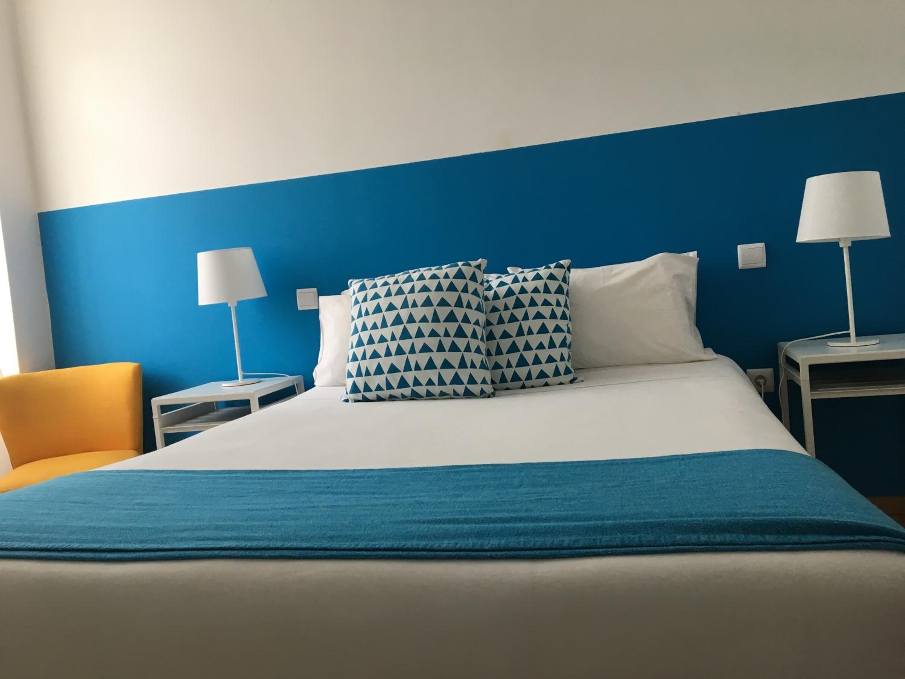 A hotel room with a bed, two bedside tables, two lamps, a blue wall, and a yellow chair.