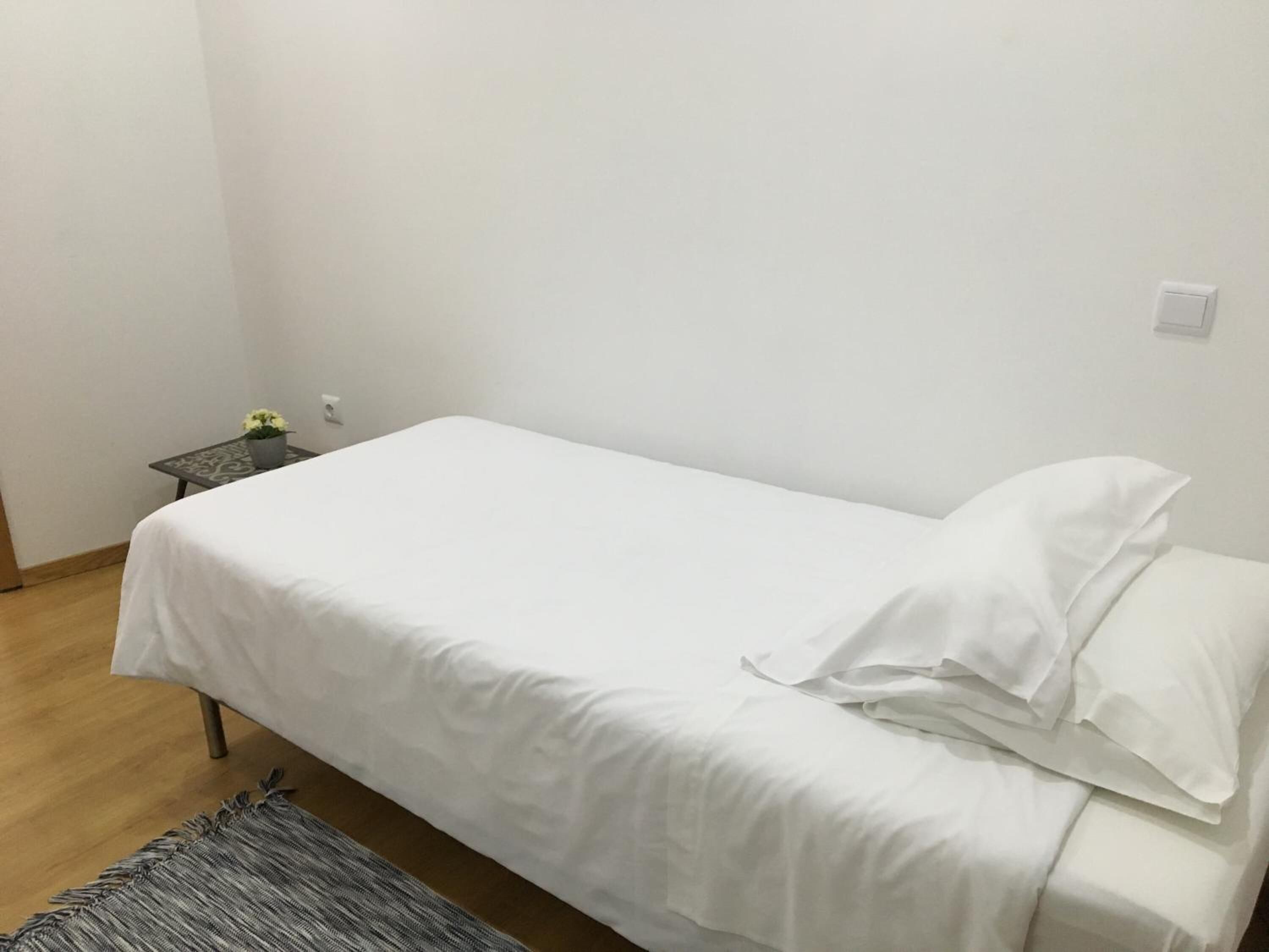 Ein Einzelbett mit weißer Bettwäsche in einem Zimmer mit kleinem Tisch und Topfpflanze.