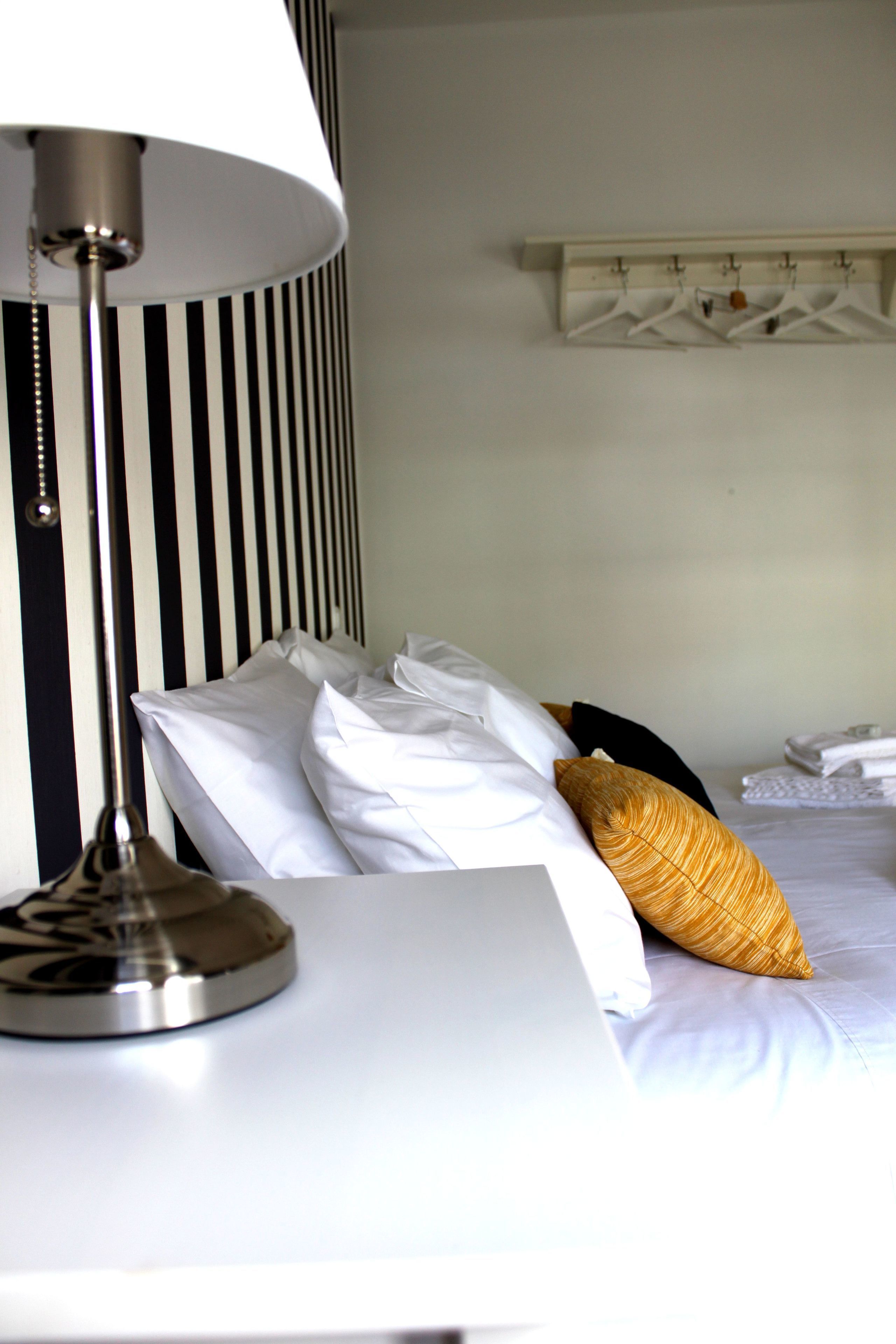 Habitació d'hotel amb llit, roba de llit blanca, paret rayada, perguam per a abrics i una làmpada.