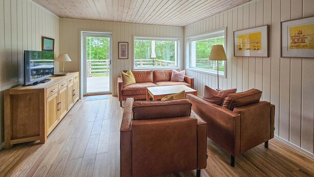 A cozy living room with a brown leather sofa set, a wooden TV stand, and a large window offering a view of the outdoors. - Nice home in Vesterøy with WiFi (Vesterøy)