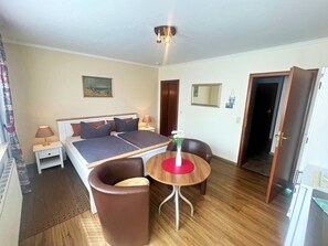 Et hotelværelse med en dobbeltseng, to sengeborde, et lille bord med en vase, en brun lænestol og trægulv.