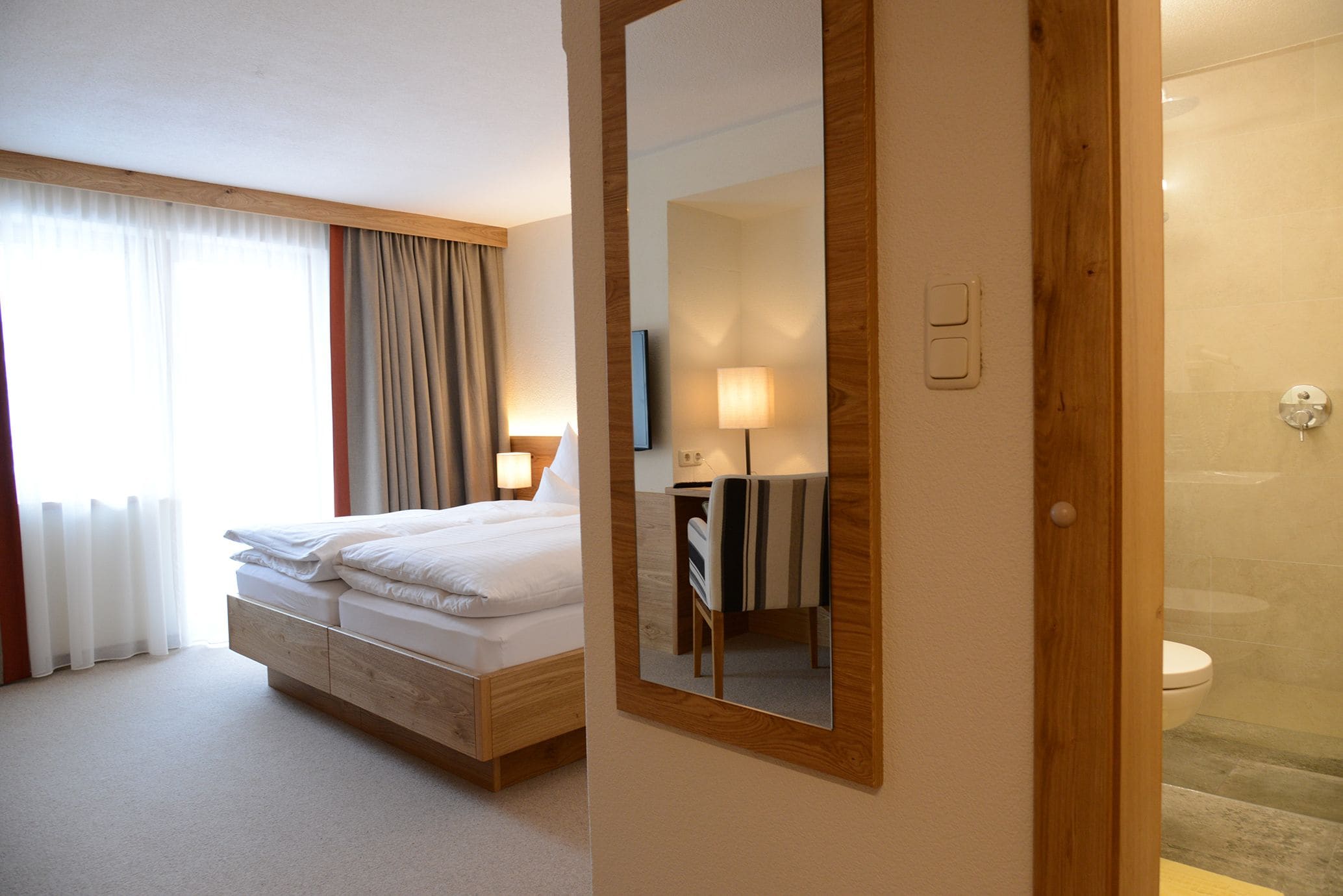 A hotel room with a bed, a mirror, a bedside lamp, and a shower area.