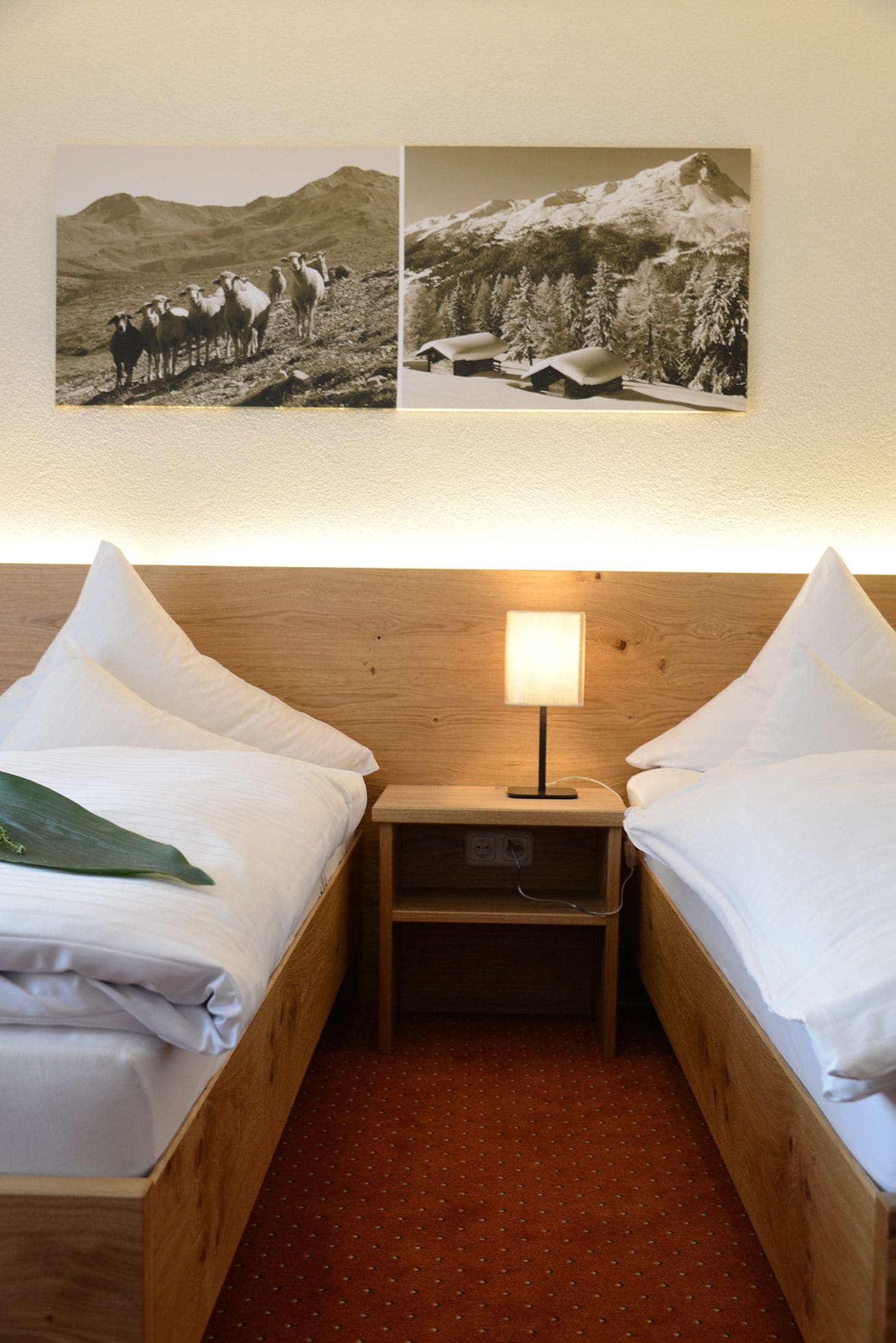 A hotel room with two wooden beds, a nightstand with a lamp, and two framed pictures on the wall.