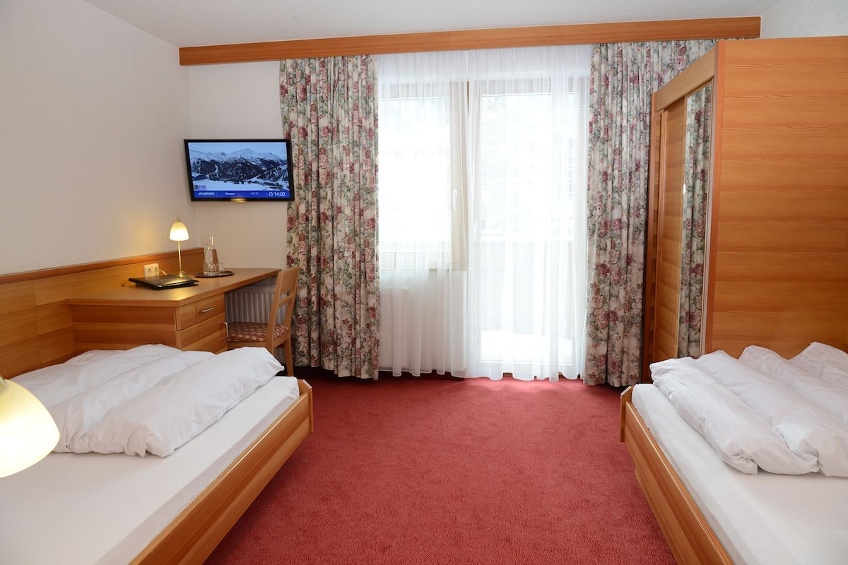 A hotel room with two beds, a television on the wall, a desk with a lamp, and a window with floral curtains.