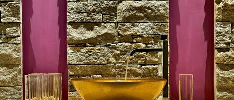 A gold bowl sink with water running, flanked by two tall glasses and a faucet.