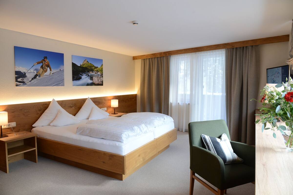 A modern hotel room with a wooden bed, two bedside tables with lamps, a green armchair, and a large window with curtains.