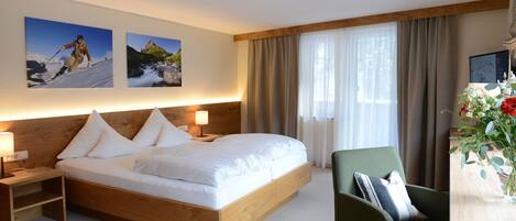 A modern hotel room with a wooden bed, two bedside tables with lamps, a green armchair, and a large window with curtains.