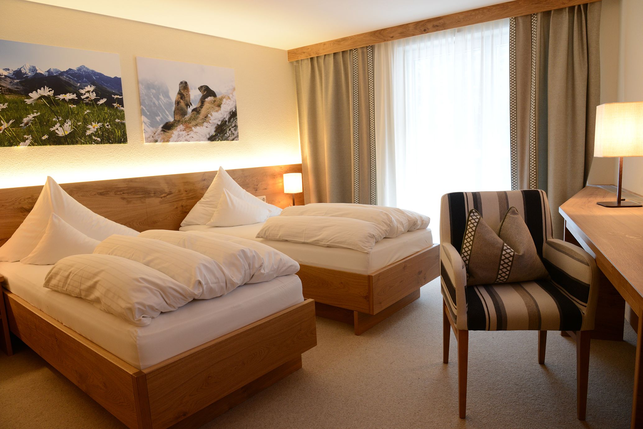 Een hotelkamer met twee houten bedden, een gestreept fauteuil, een bureau met een lamp en twee ingelijste foto's aan de muur.