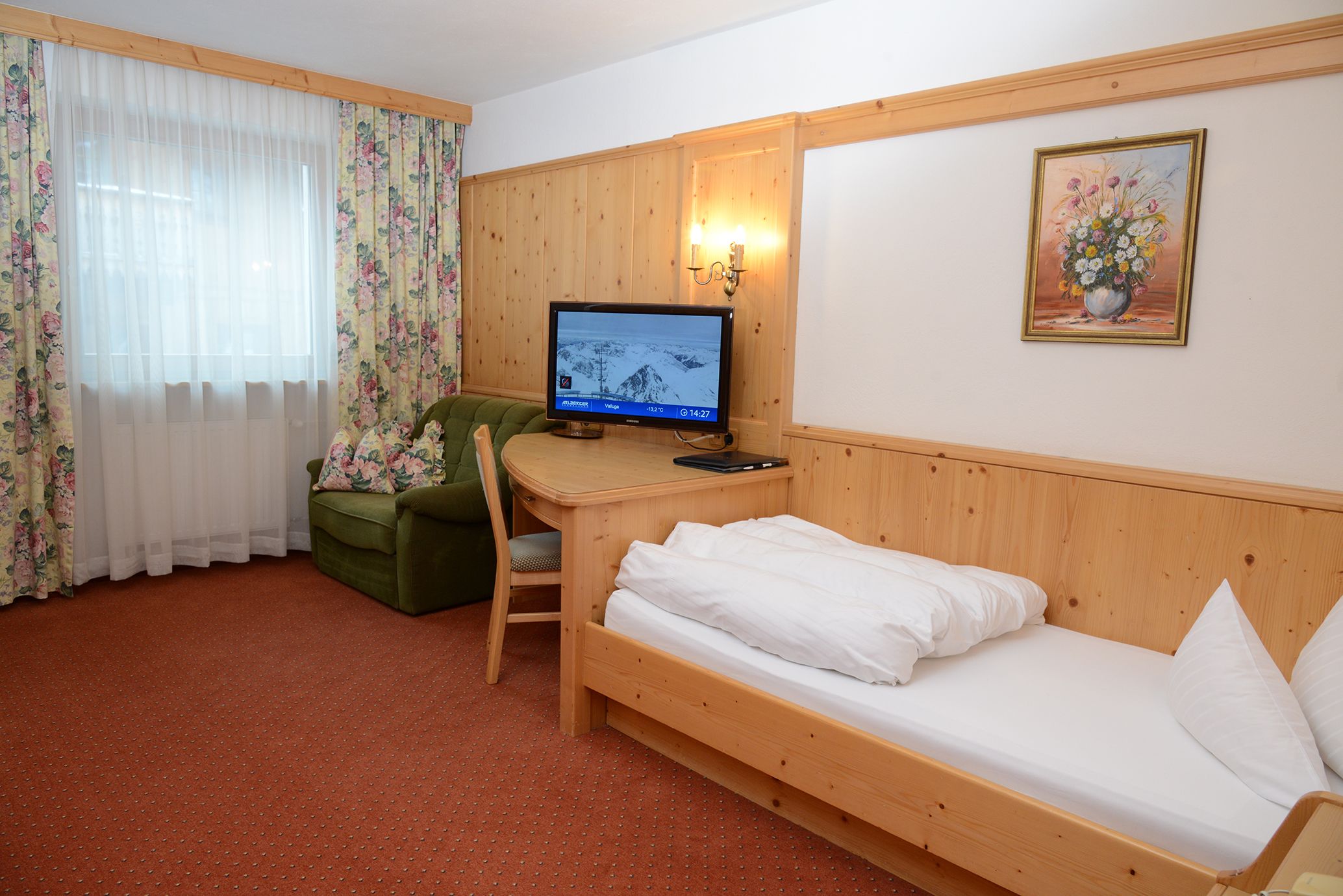 A room with a wooden bed, a desk with a computer, a green chair, and a painting on the wall.