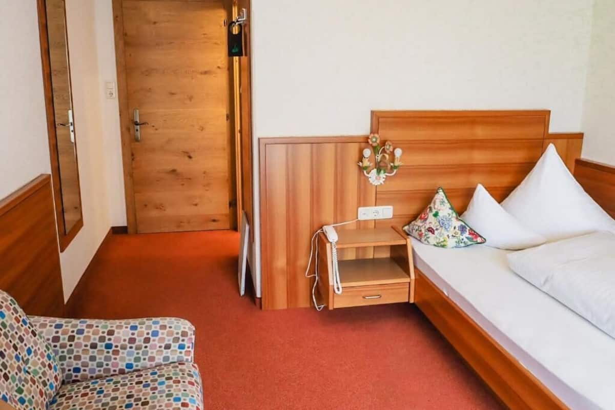 A hotel room with a wooden bed, a small bedside table with a phone, a floral pillow, and a patterned armchair.