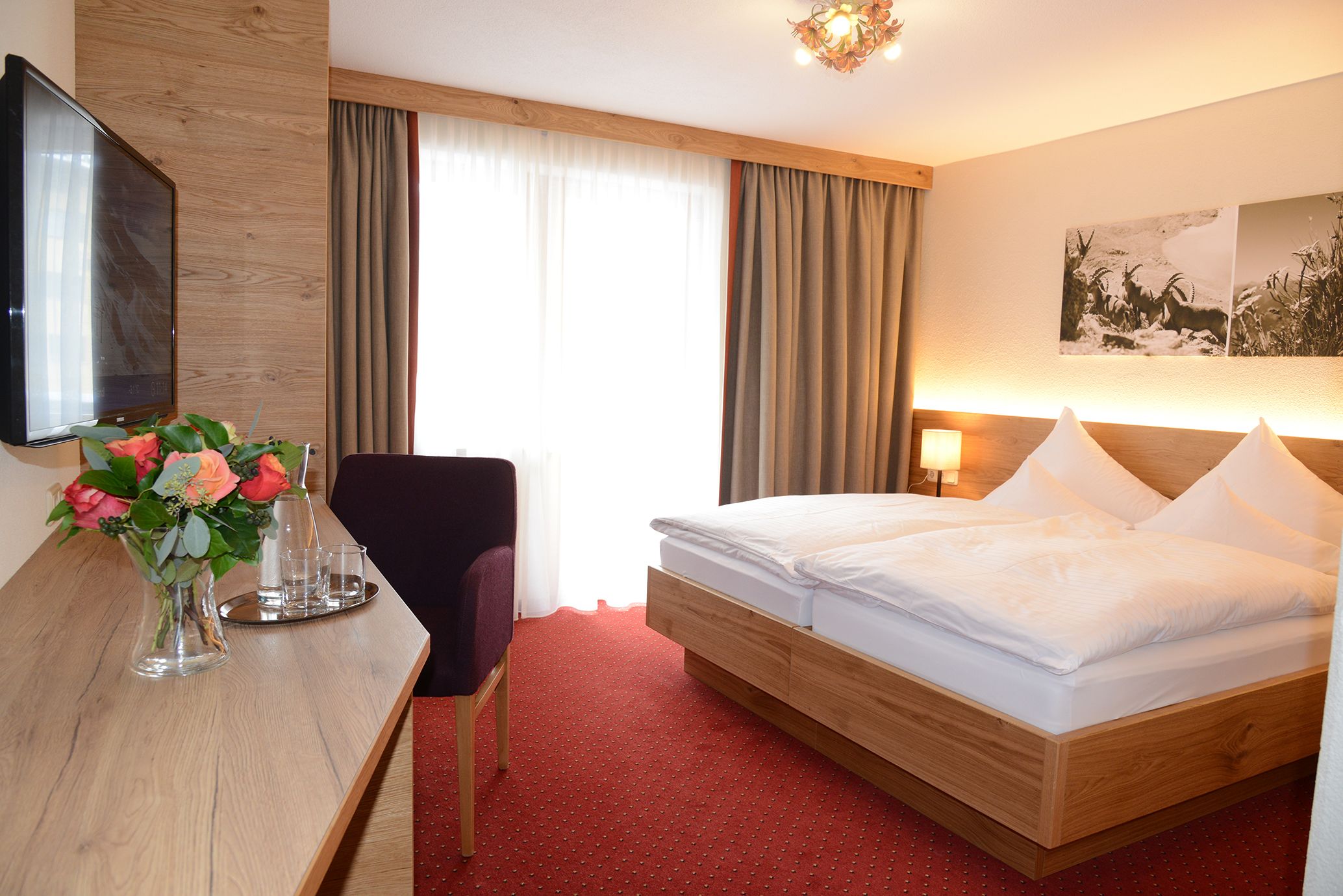 A hotel room with a wooden bed, a desk with a vase of flowers, a chair, and a television.
