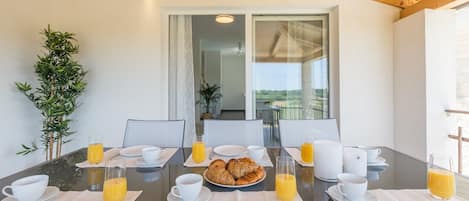 A breakfast table set with plates, cups, and glasses of orange juice, with a view of the outdoors through a large window.