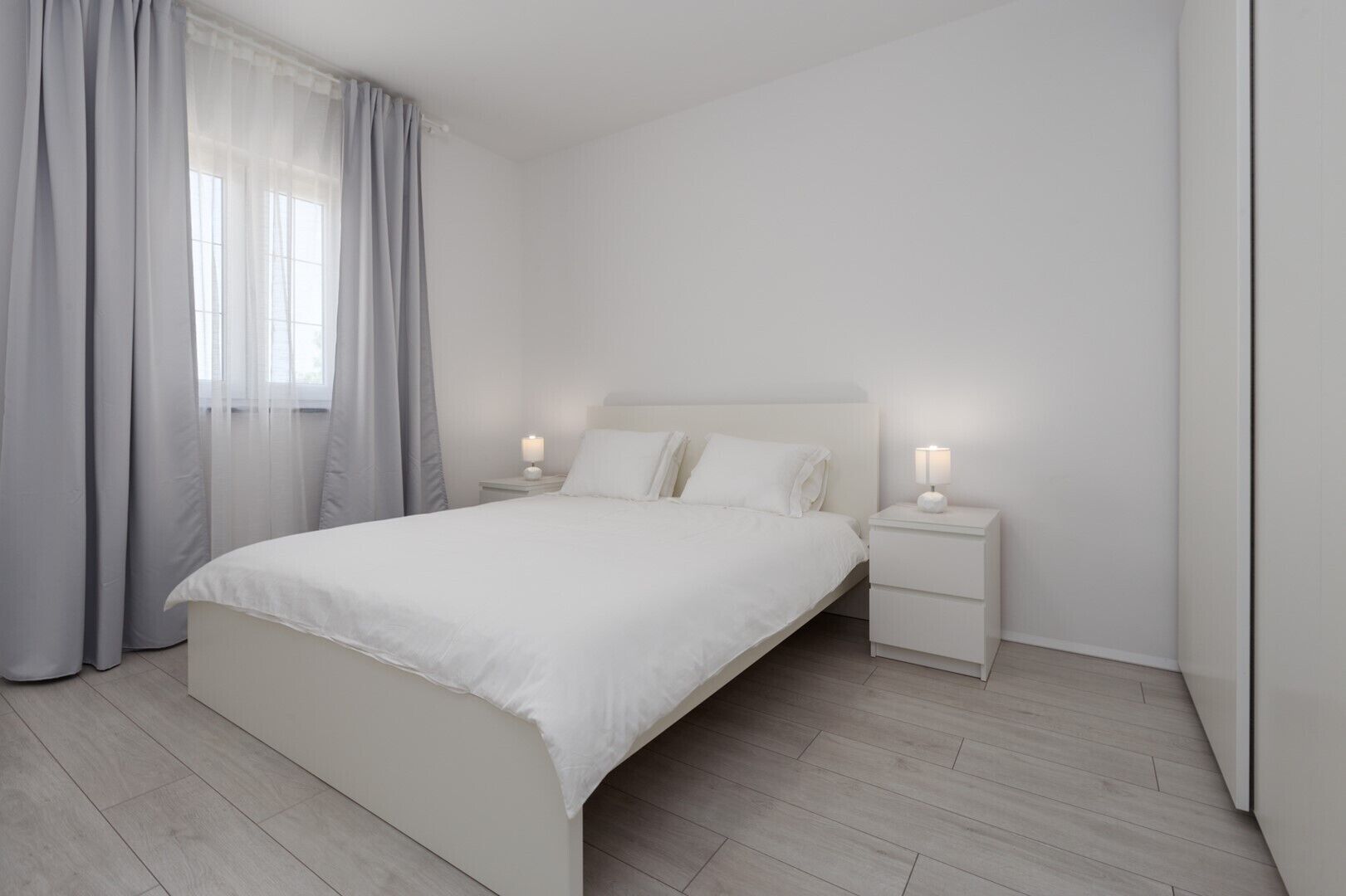 Un dormitorio minimalista con una cama grande, dos mesitas de noche con lámparas y un ventanal con cortinas traslúcidas.