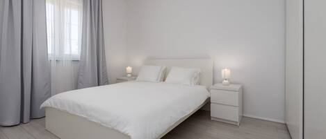 Un dormitorio minimalista con una cama grande, dos mesitas de noche con lámparas y un ventanal con cortinas traslúcidas.