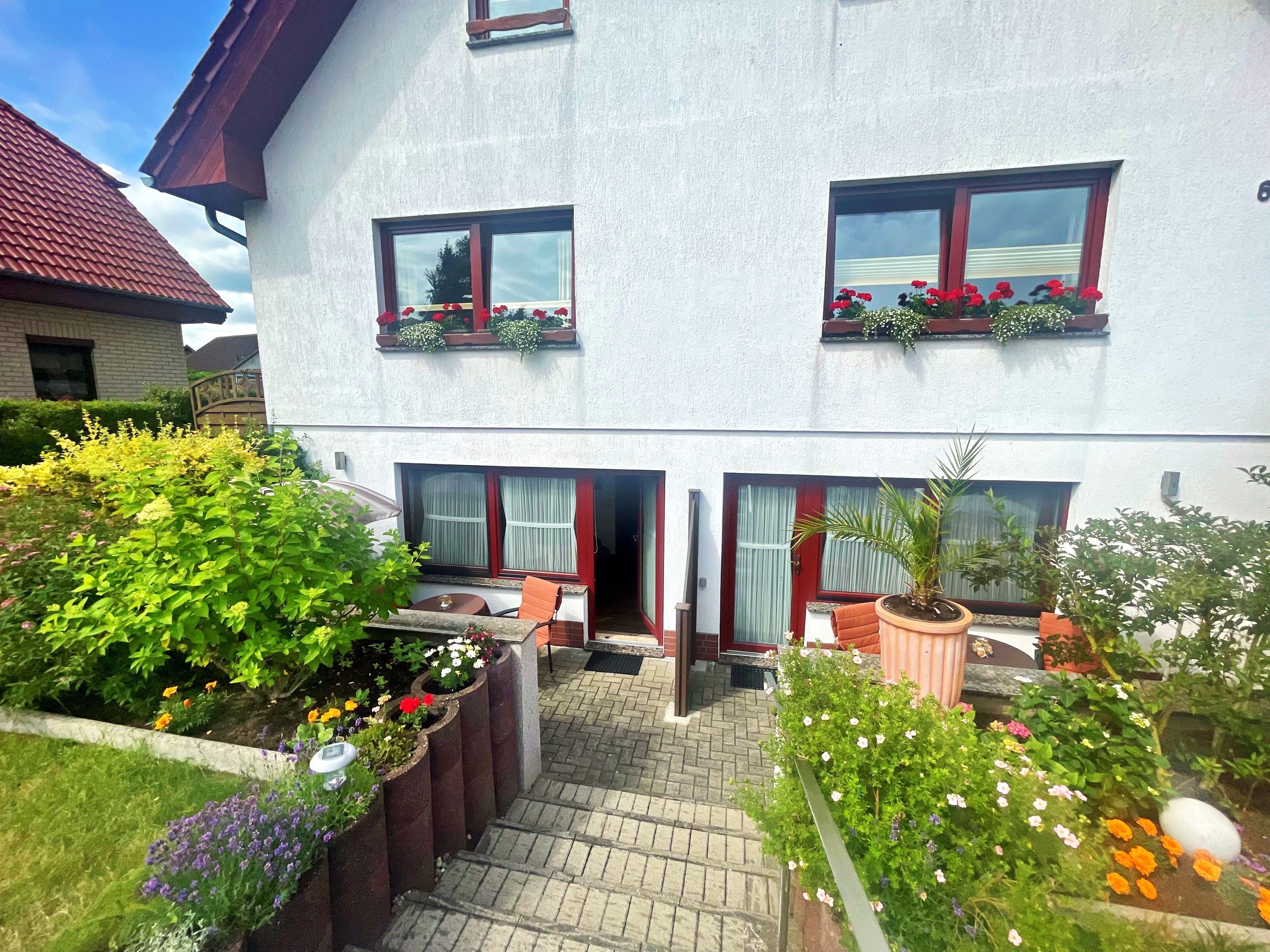 Un edificio de dos pisos con fachada blanca, múltiples ventanas con jardineras y un área de entrada pavimentada con macetas y un pequeño espacio para sentarse.