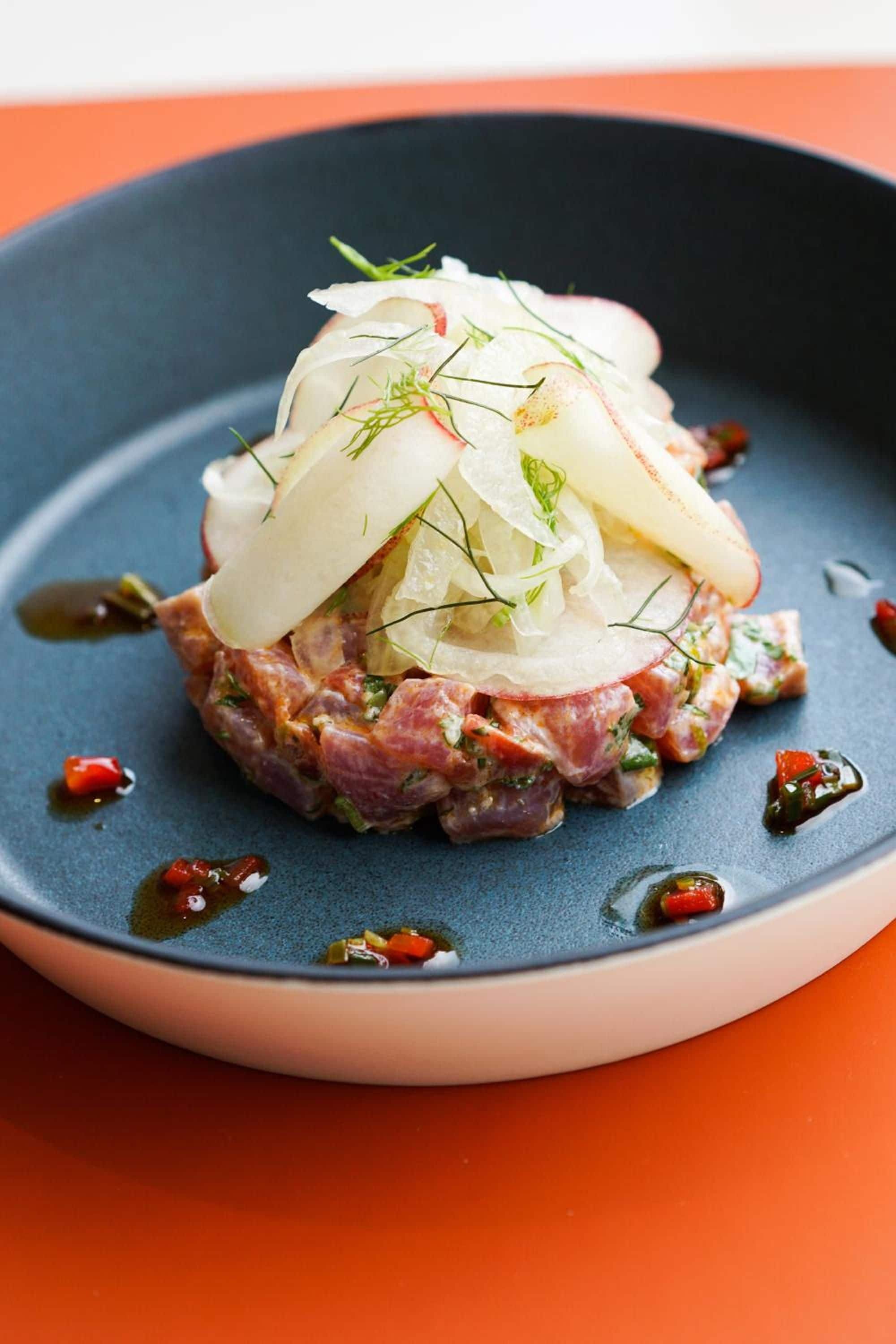 A dish with a base of minced meat, topped with thinly sliced onions and herbs, served on a dark plate with a blue rim.