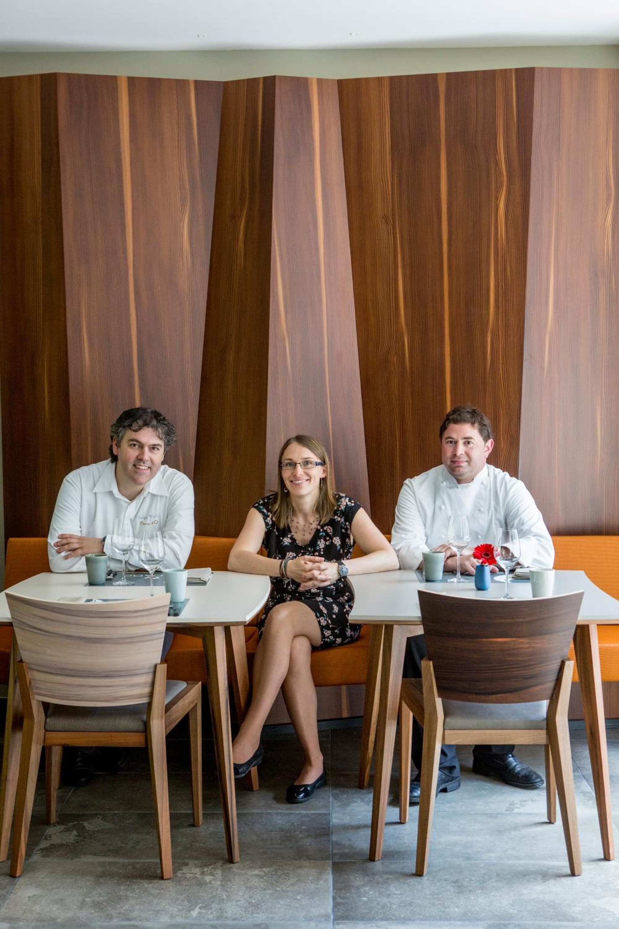 Drie personen zitten aan een tafel, met een houten wand op de achtergrond.