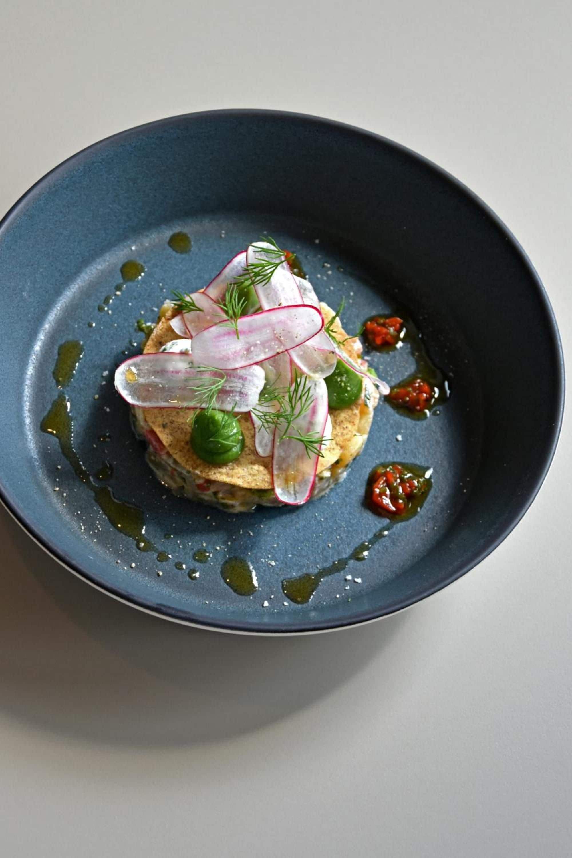 A plate with a layered dish, garnished with radishes and herbs, and surrounded by olive oil and a red sauce.
