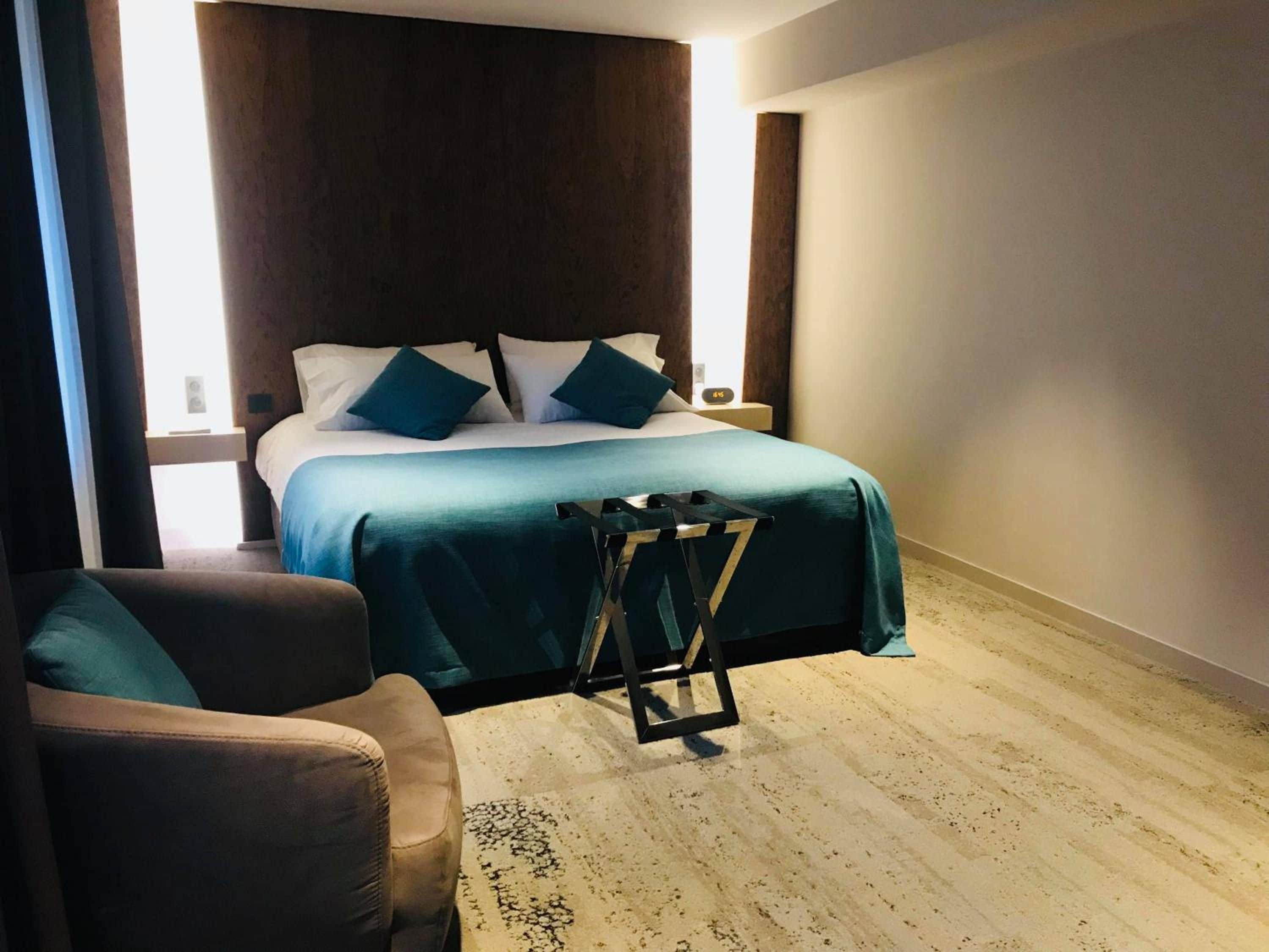 Quarto de hotel com uma cama, uma cadeira e uma mesinha.