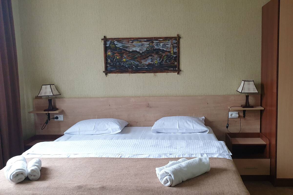 A neatly made bed with two pillows and two rolled towels, a wooden headboard, a painting on the wall, two bedside tables with lamps, and a wardrobe.