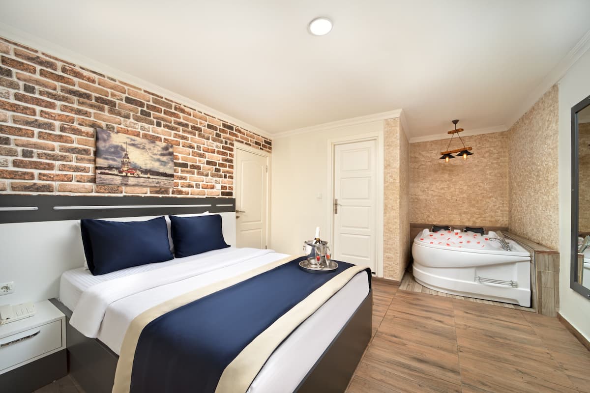 A hotel room with a brick wall, a large bed with navy blue bedding, a white door, a wooden floor, and a white bathtub with red flowers.