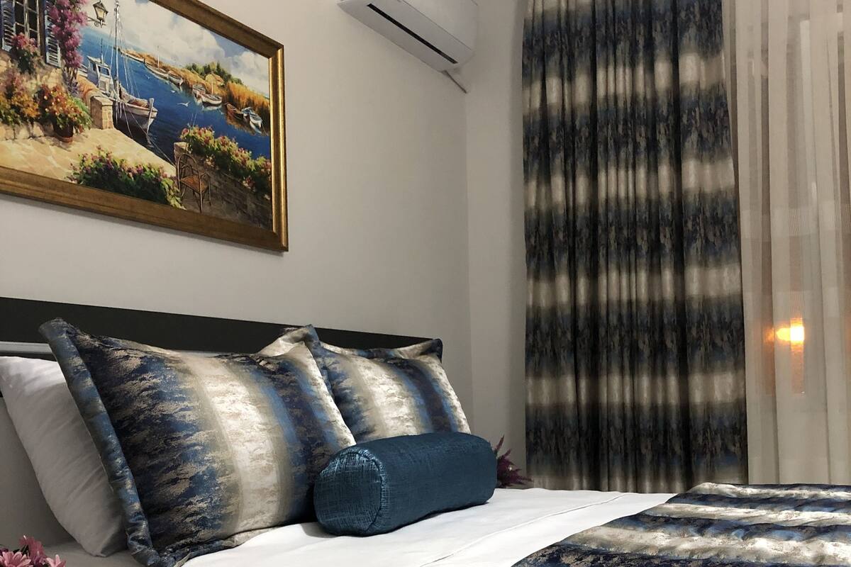 A hotel room with a bed, a painting, an air conditioner, a curtain, and a coffee cup on a table.
