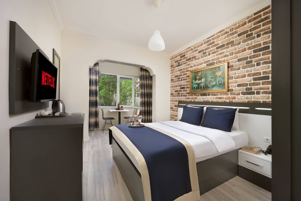 A modern hotel room with a brick wall, a bed with a headboard, a TV, a desk with a chair, and a window with curtains.