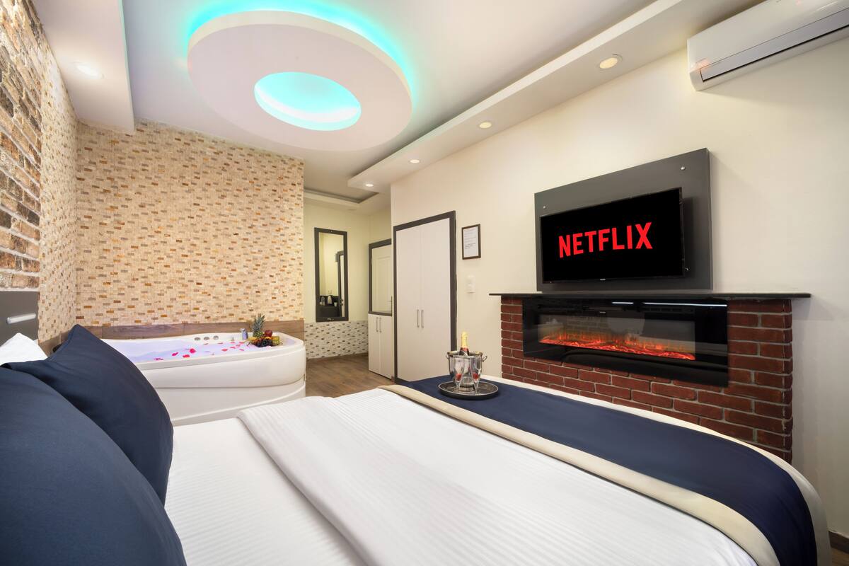 A modern hotel room with a brick fireplace, a large bed, a bathtub, and a flat-screen TV mounted above the fireplace.