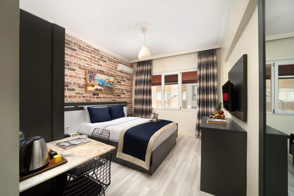 A compact hotel room with a bed, a desk with a kettle, a TV, and a brick wall with a painting.