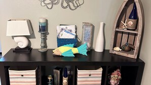 Una consola negra con cajones de almacenamiento, un termómetro de pared, un pulpo decorativo, un cuadro enmarco, una pequeña estantería con una concha marina y una estrella de mar, y una lámpara.