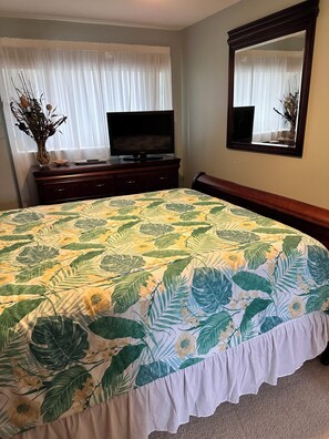 Une chambre à coucher avec un lit, un téléviseur posé sur une commode, un miroir au mur et une fenêtre avec des rideaux.