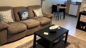 Una sala de estar con sofá, mesa de centro y un comedor con mesa y sillas. Hay un ventilador de techo, una lámpara y cuadros en la pared.