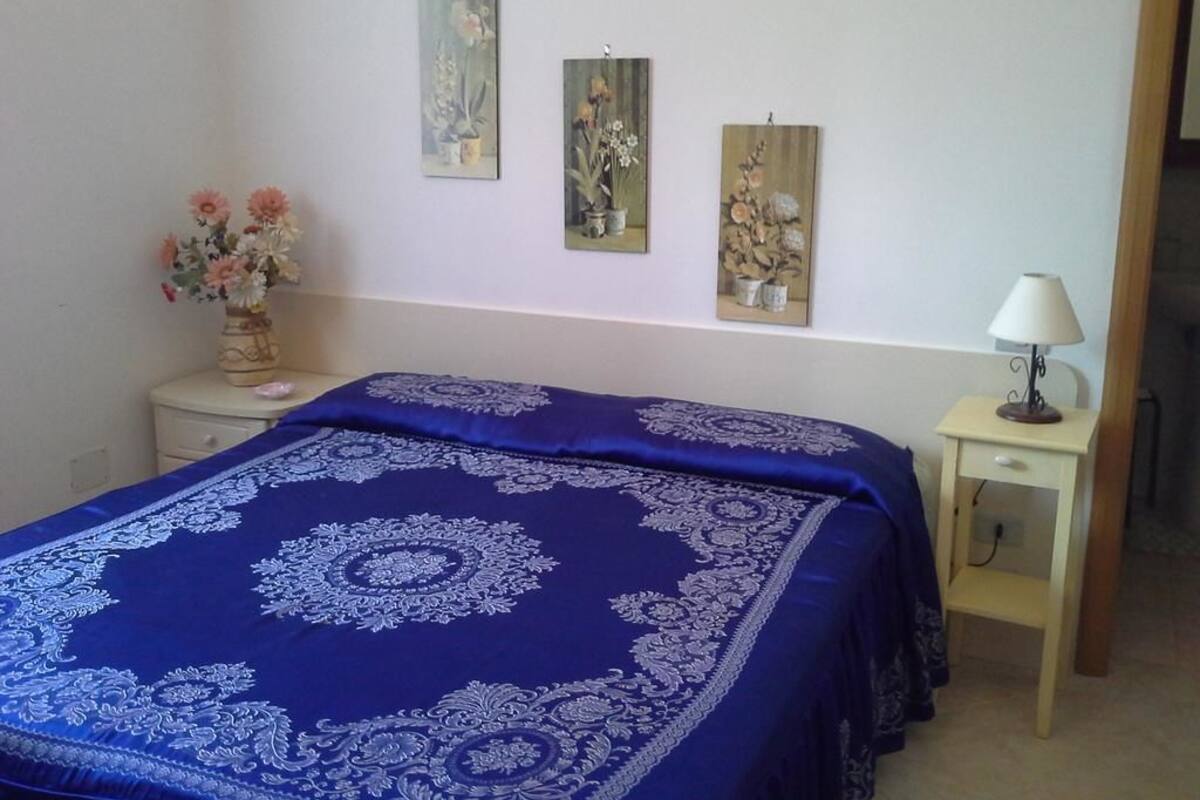 Un dormitori amb un llit gran cobert amb un edredó estampat blau i blanc, una taula de nit amb una làmpada i tres quadres penjats a la paret.
