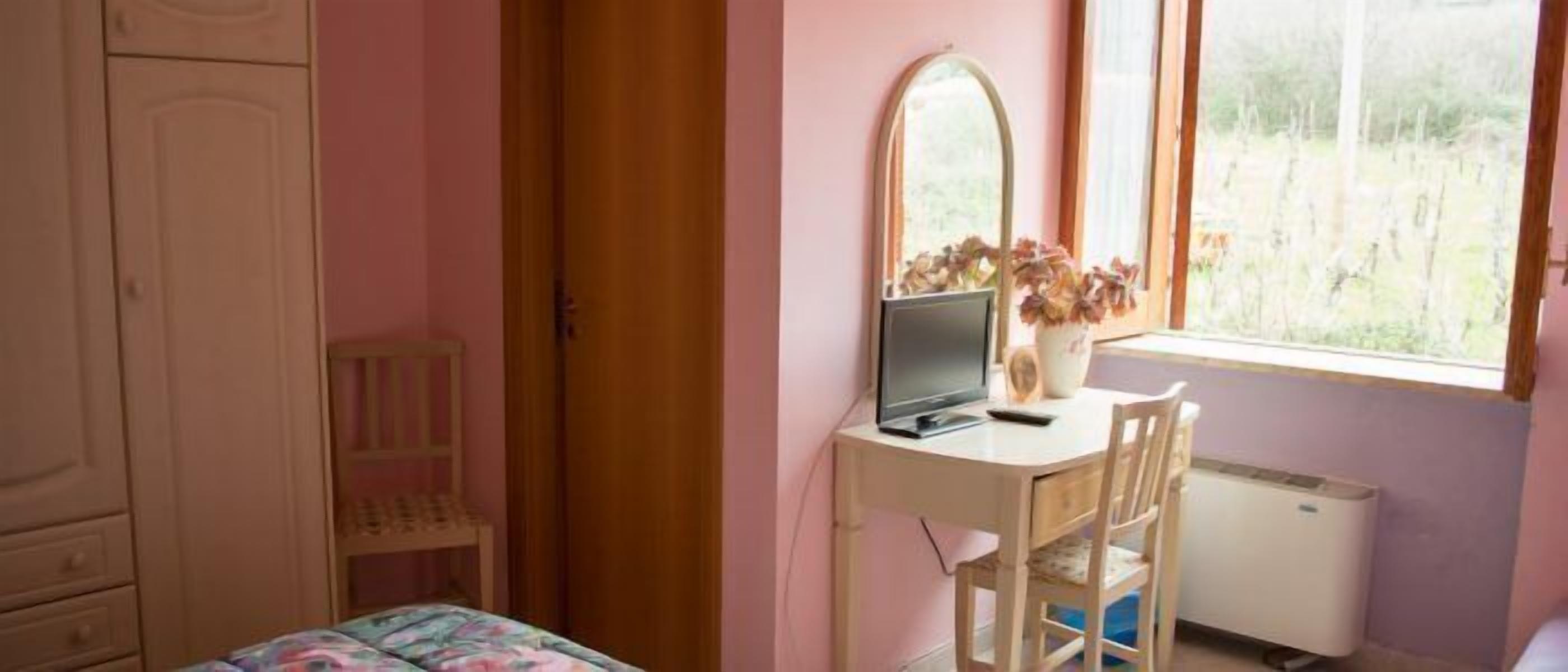 Una camera da letto con un letto, una scrivania con un computer, una sedia, una finestra vista su del verde e un armadio.