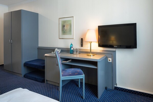 A hotel room with a bed, a desk with a chair, a television, a lamp, and a remote control.