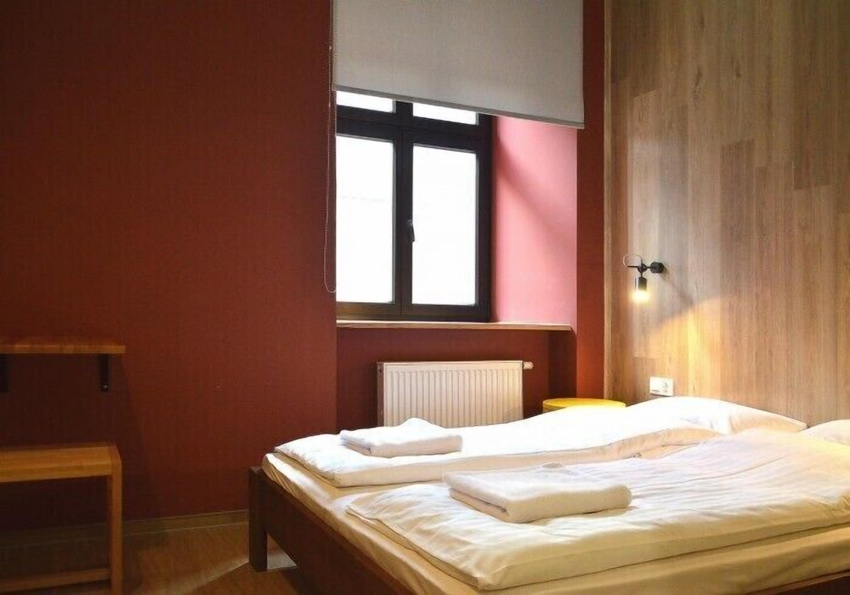 A bedroom with two beds, a window with blinds, a radiator, and a wooden wall.